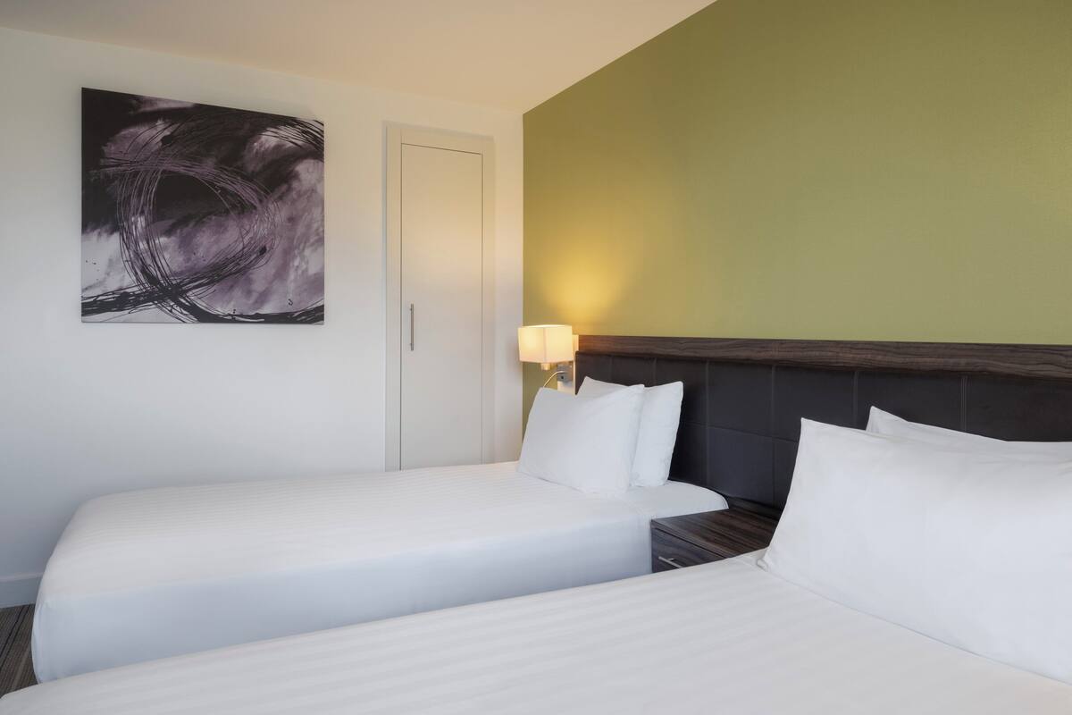 A hotel room with two beds, a nightstand, a wall-mounted lamp, and a large abstract painting.