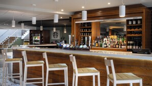 A bar area with wooden bar stools, a counter with a cash register, and shelves stocked with bottles. - Holiday Inn Huntingdon - Racecourse by IHG (Huntingdon)