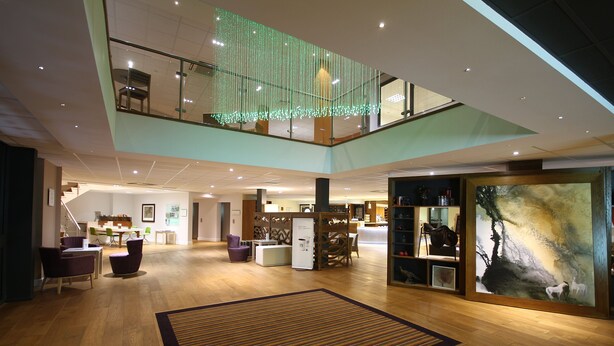 A spacious lobby with a high ceiling, a central staircase, and a large artwork on the right wall. - Holiday Inn Huntingdon - Racecourse by IHG (Huntingdon)