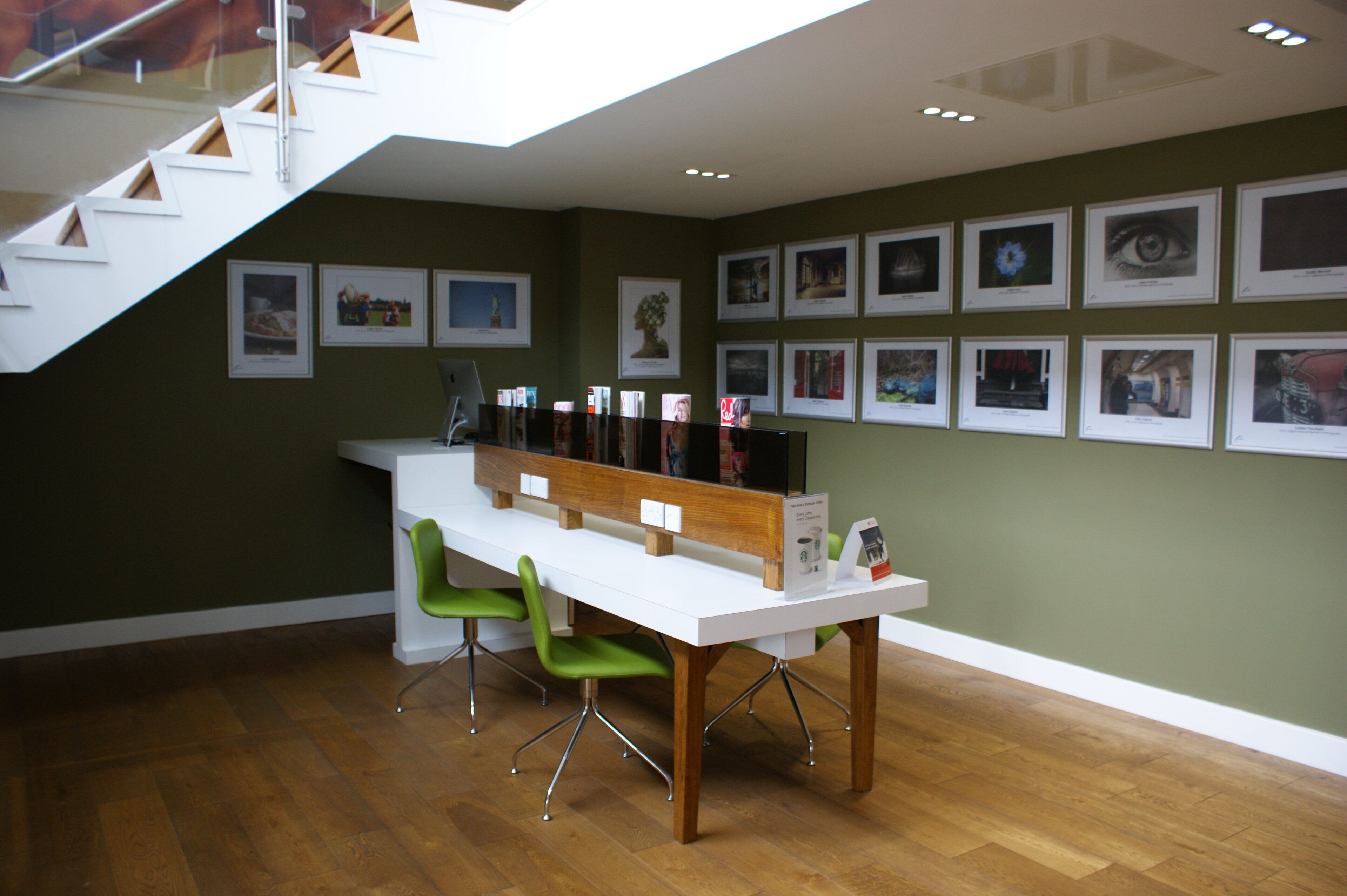 Un intérieur moderne avec un escalier, une table blanche et des chaises vertes, et un mur décoré de tableaux encadrés.