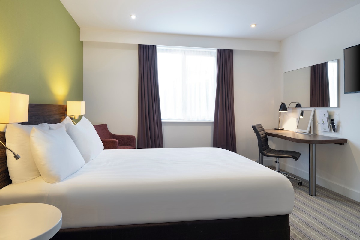 A hotel room with a large bed, a desk with a chair, a desk lamp, a flat-screen TV, and a window with curtains.