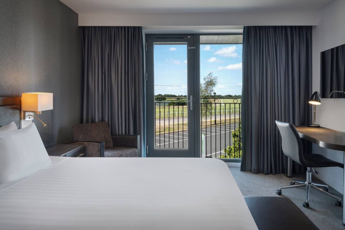 A hotel room with a bed, a desk, a chair, a lamp, and a view of a parking lot through a glass door.