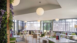 A spacious dining area with wooden tables and green chairs, large windows, and hanging pendant lights. - Holiday Inn Huntingdon - Racecourse by IHG (Huntingdon)