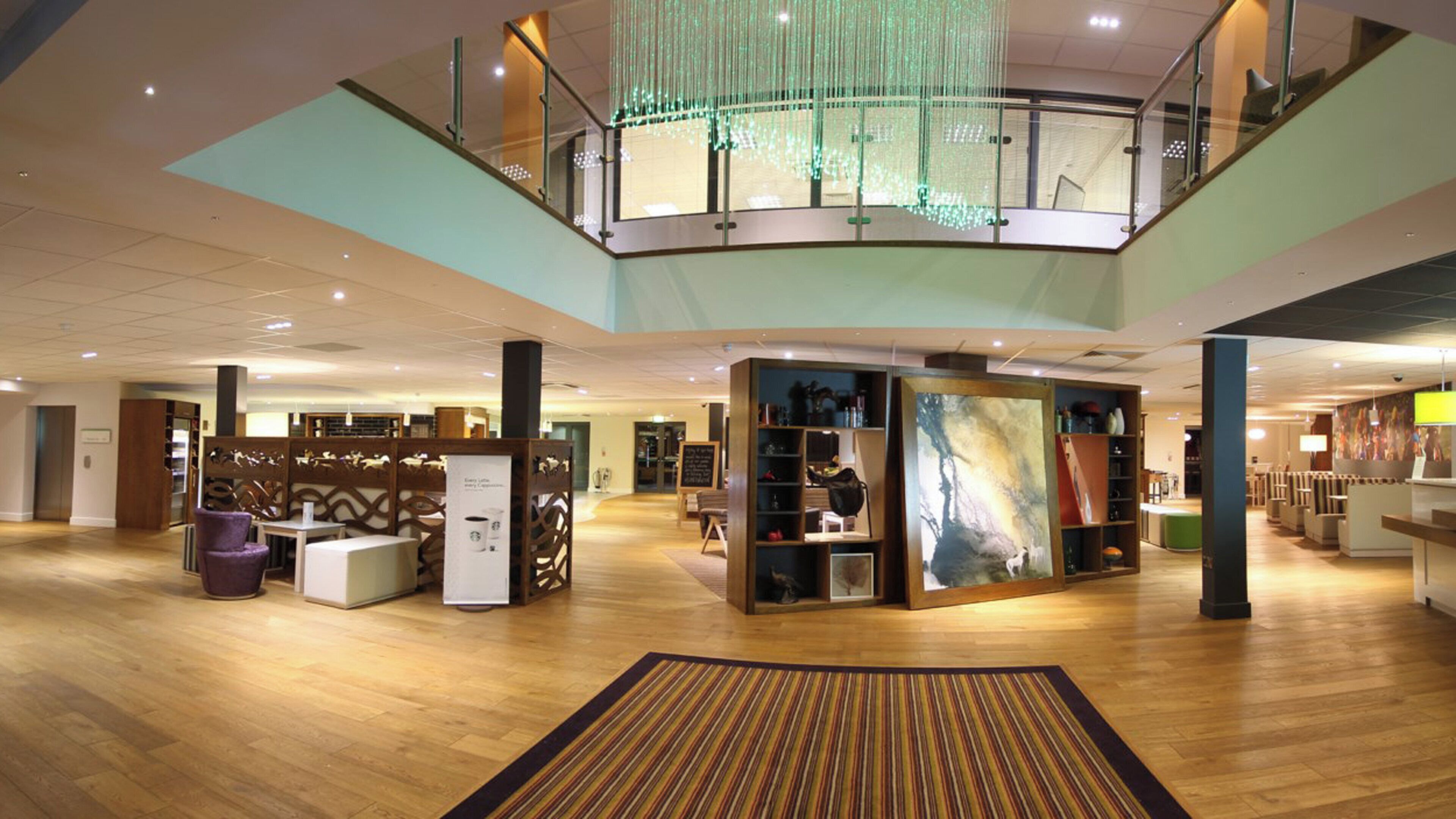 A spacious lobby with a wooden floor, a central seating area, and a mezzanine level with a glass railing.