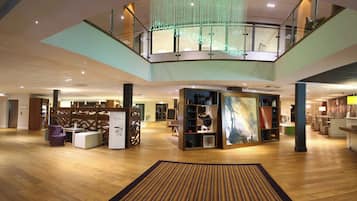 A spacious lobby with a wooden floor, a central seating area, and a mezzanine level with a glass railing.