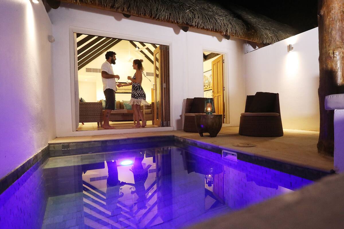 Ein Paar steht an einem Pool mit einem Reetdach und blickt in einen Raum mit einem Bett und einem Nachttisch.