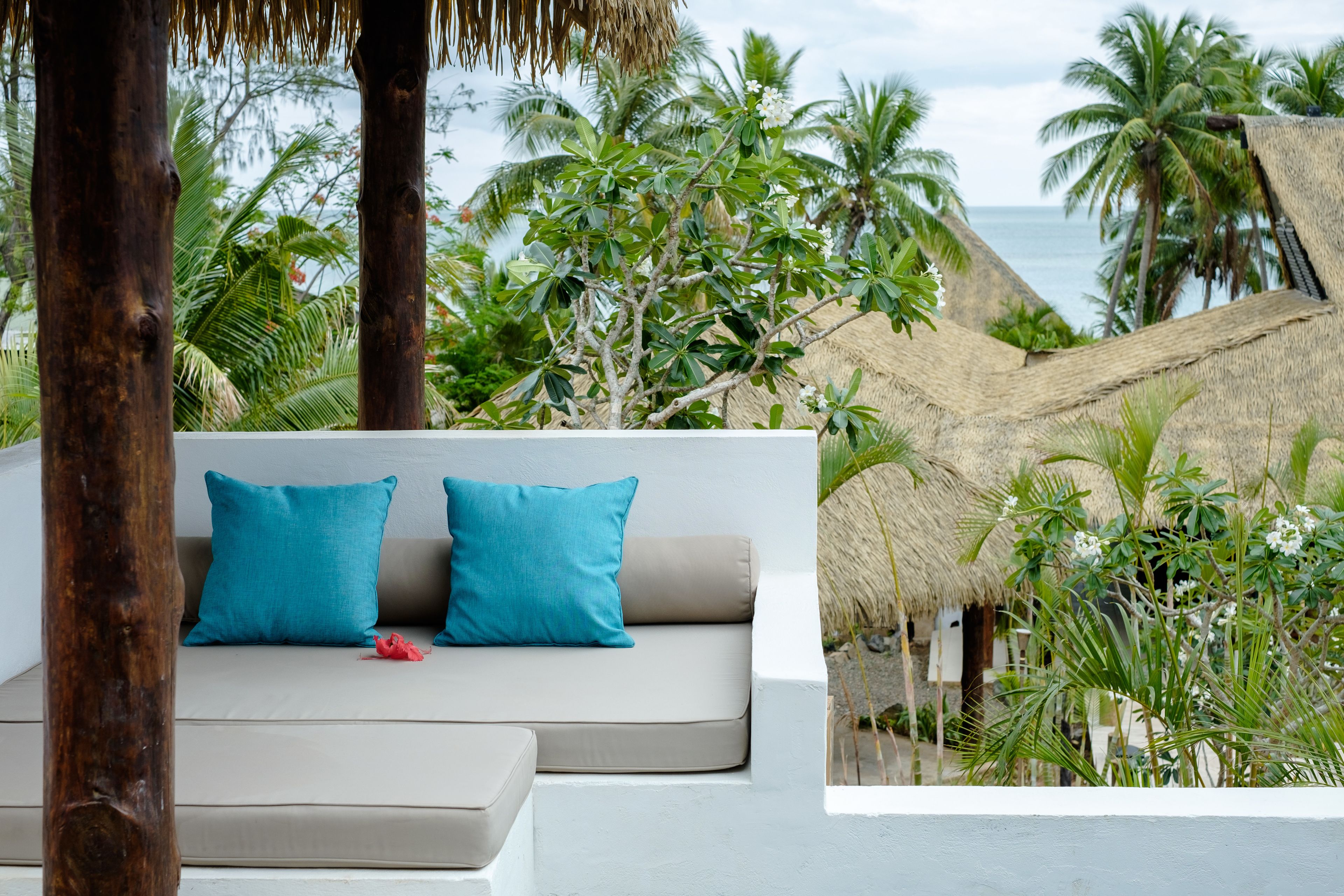 A modern outdoor seating area with a sofa and cushions, surrounded by tropical vegetation and a view of the ocean.