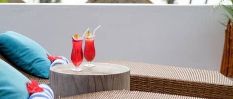 A wicker lounge chair with a wooden table, two red drinks with straws, and a rolled towel with a blue and white striped towel and a red flower.
