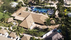 Luchtbeeld van een resort met gebouwen met rieten daken, een zwembad en palmbomen.