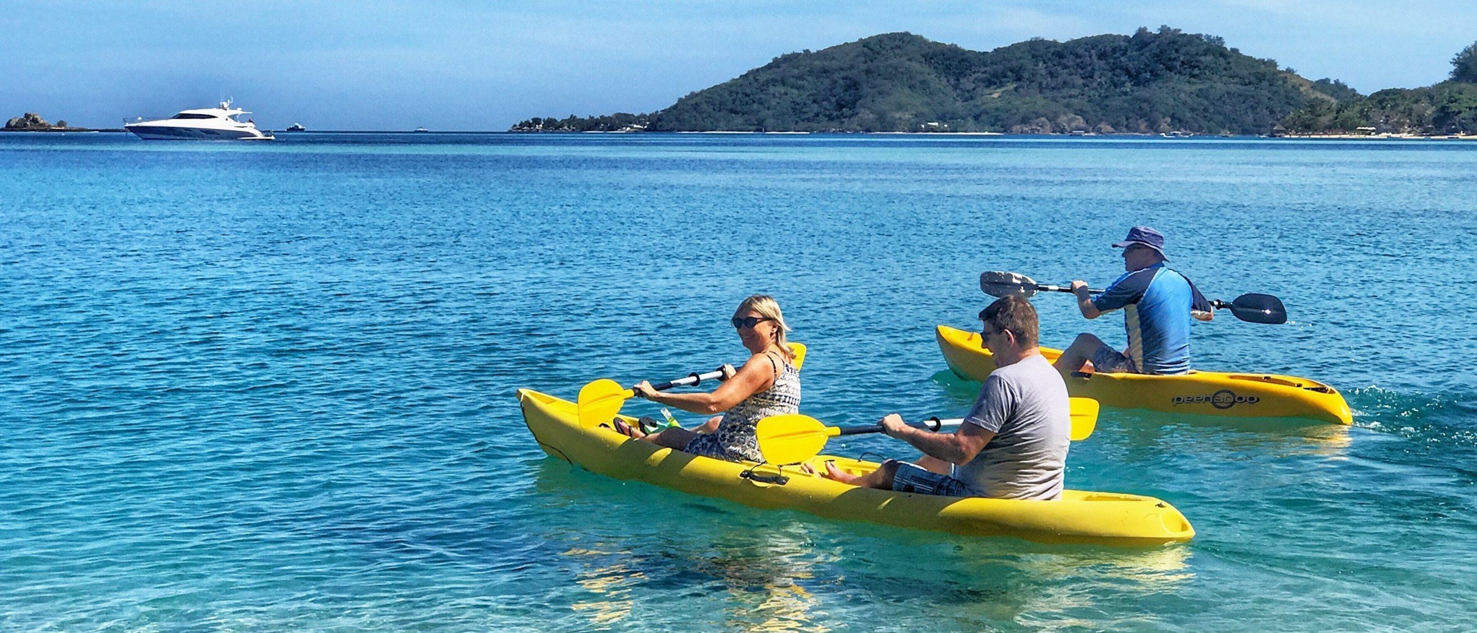 Dos personas haciendo kayak en un mar tranquilo, con un yate al fondo y una isla boscosa.