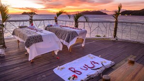 Zonsondergang uitzicht vanaf een houten terras met twee bedden, palmbomen en een tafel met een naam op een mat.