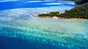 Vue aérienne d’une zone côtière avec des eaux turquoise limpides, une petite île sur laquelle se trouve un bâtiment, et une végétation luxuriante.