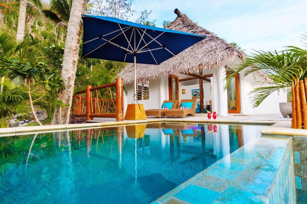 Ein Poolbereich mit einer strohgedeckten Struktur, Liegestühlen und einem Pool, der die Umgebung spiegelt.