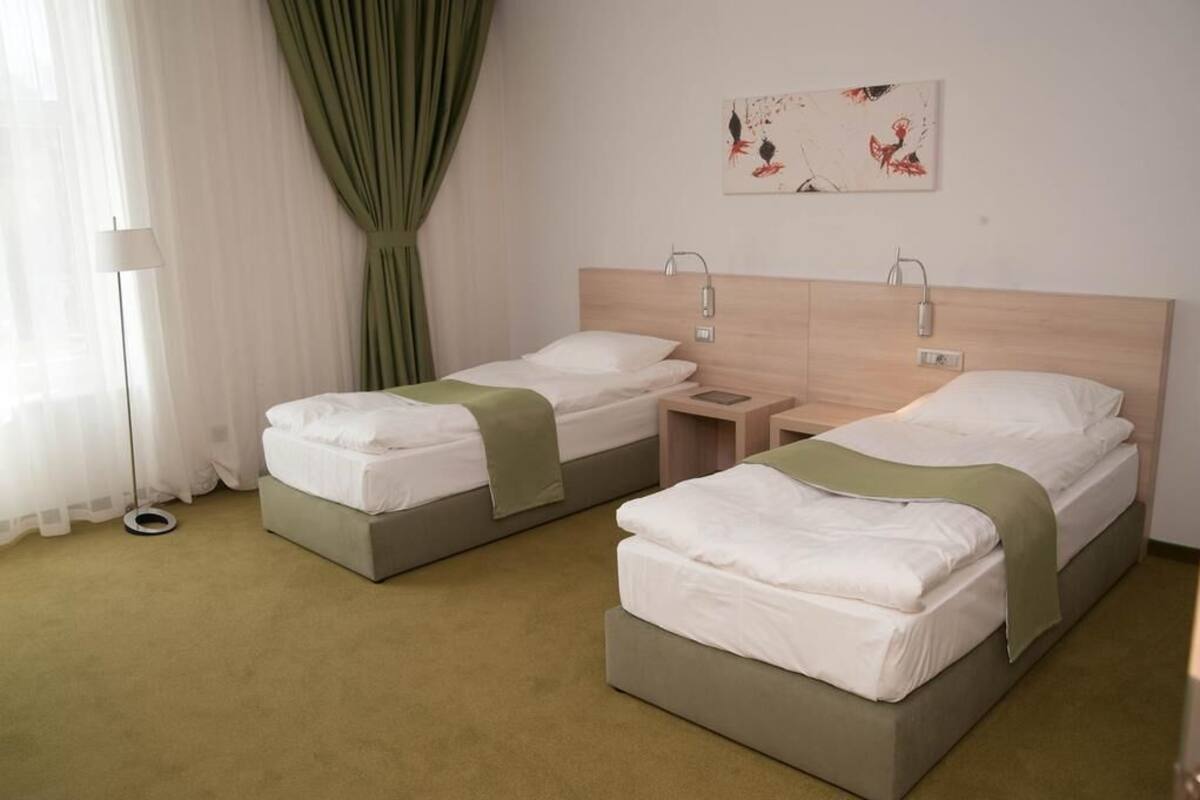 A hotel room with two single beds, a bedside table with a lamp, a wall-mounted artwork, and a floor lamp.
