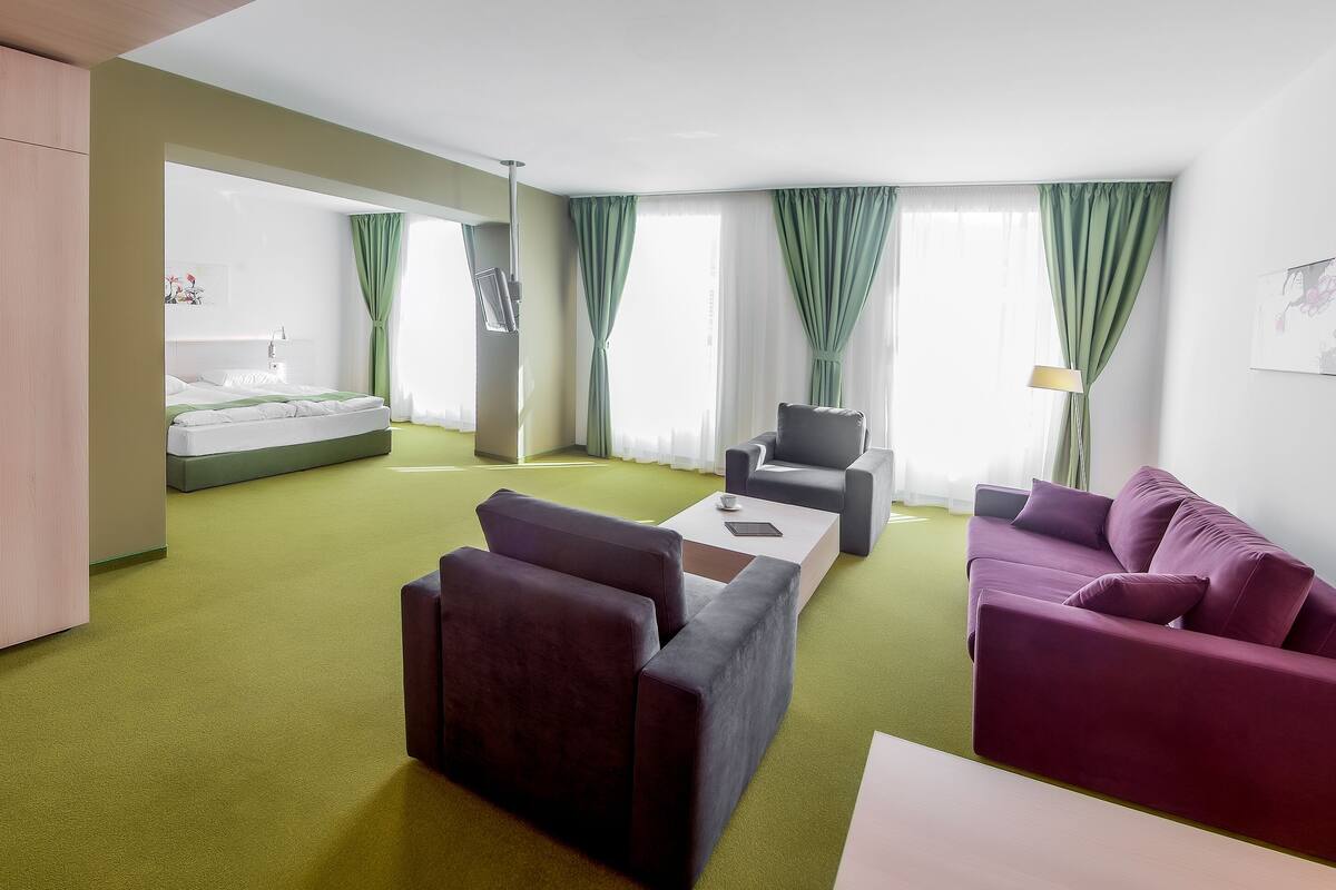 A modern hotel room with a green carpet, a bed, a sofa, a chair, and a desk.