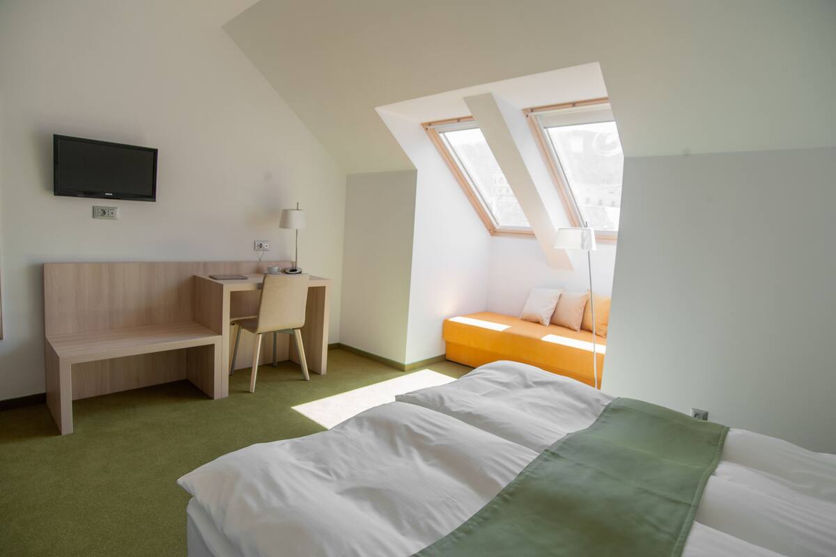 A modern hotel room with a sloped ceiling, a bed with white bedding, a desk with a chair, a television mounted on the wall, and a window providing natural light.