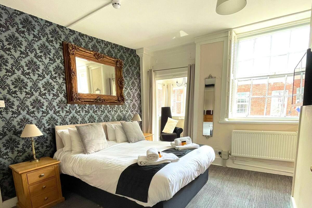 A neatly made bed with a black and white duvet, a wooden nightstand with a lamp, and a large mirror on the wall.