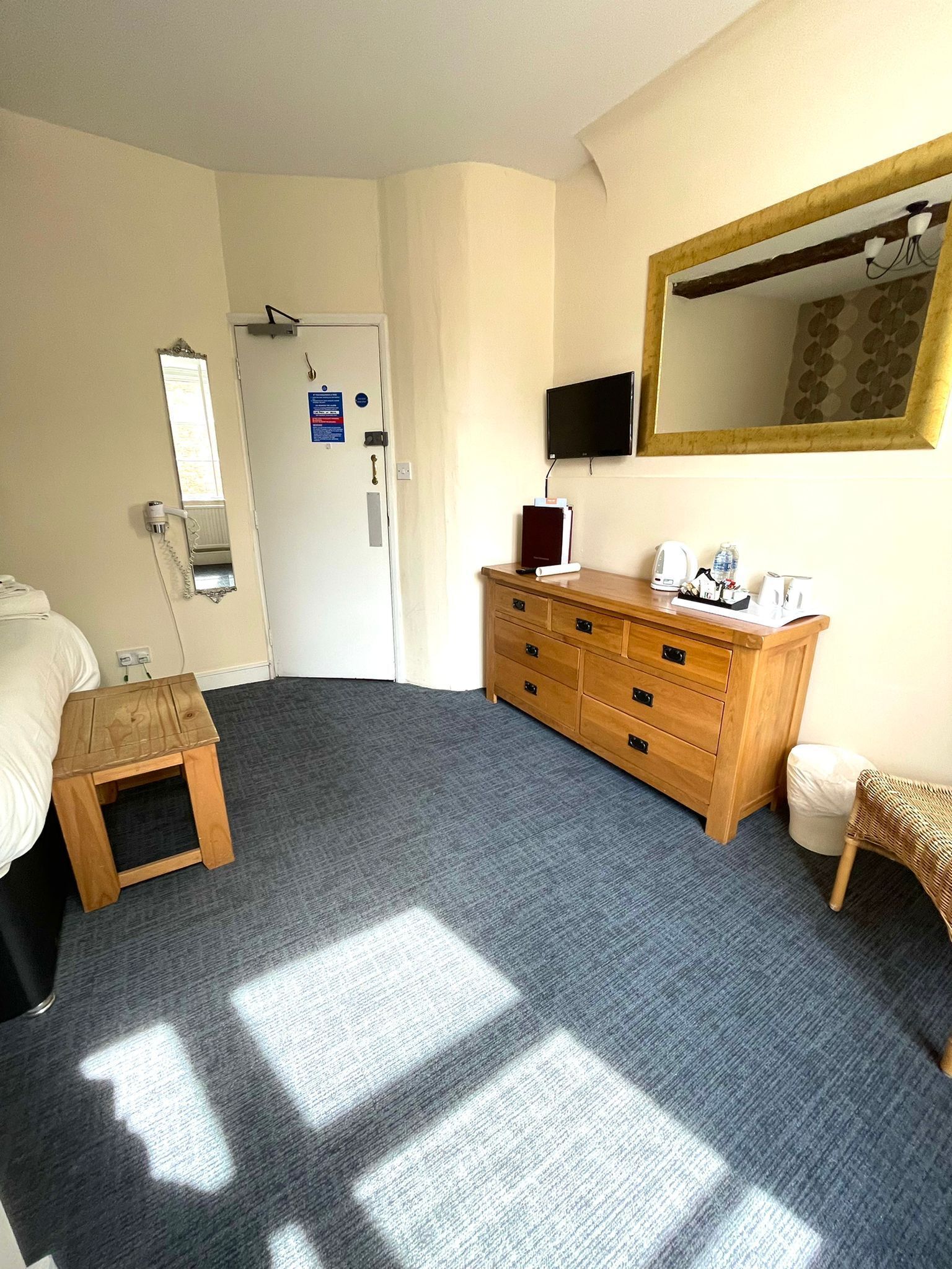 Una habitación de hotel con una cama, un aparador de madera, un taburete pequeño de madera, un cubo de basura, un espejo y un televisor fijado a la pared.