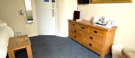 Una habitación de hotel con una cama, un aparador de madera, un taburete pequeño de madera, un cubo de basura, un espejo y un televisor fijado a la pared.
