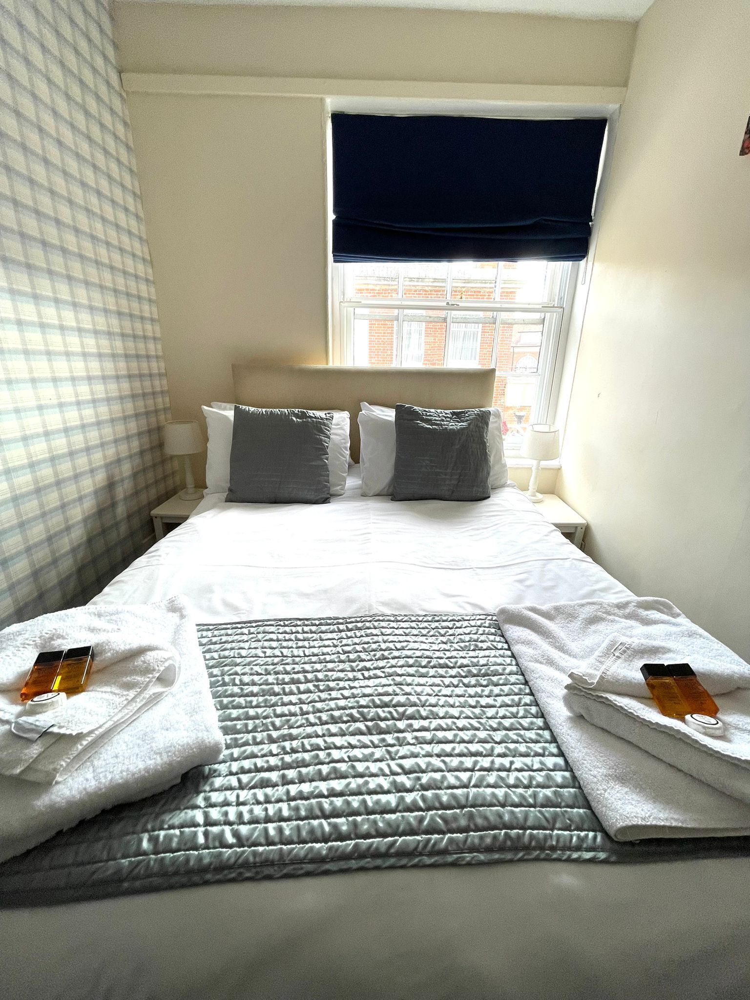 A hotel room with a neatly made bed, a window with a view of buildings, and a checkered curtain.