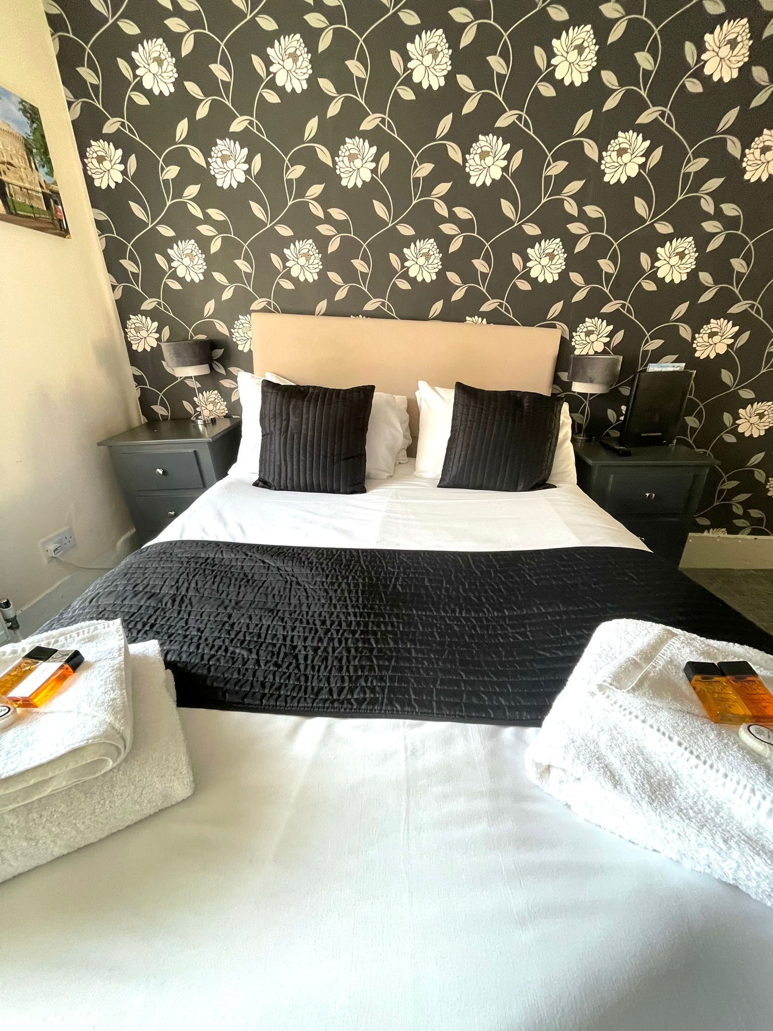 Una habitación de hotel con una cama, dos mesitas de noche, una lámpara y un papel tapiz con motivos florales.
