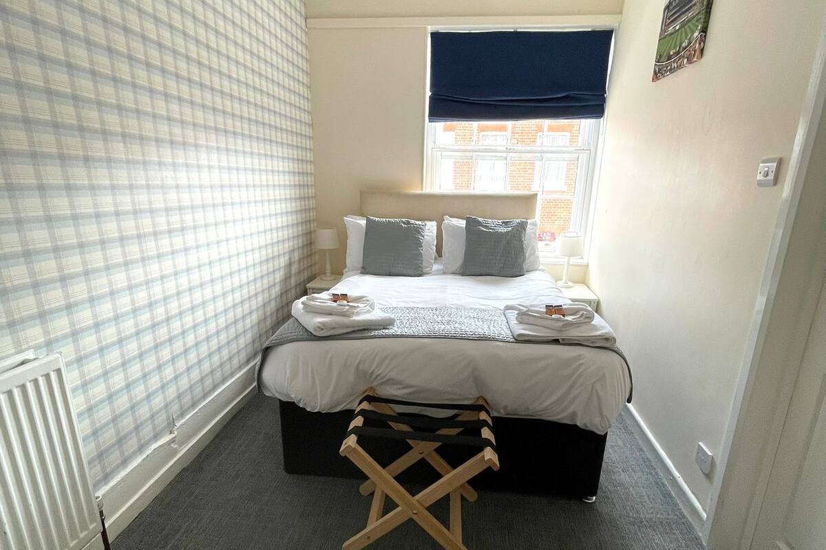 A bedroom with a bed, a radiator, a window with a blue blind, and a small wooden stool.