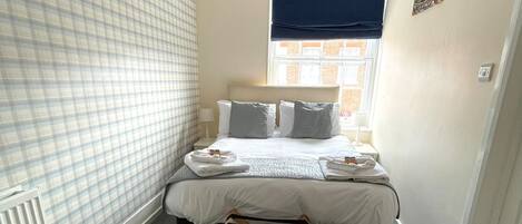 A bedroom with a bed, a radiator, a window with a blue blind, and a small wooden stool.