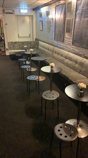 A dimly lit interior with a row of round tables and stools, a cushioned bench, and a wall with framed pictures. - The Butterfly Hotel - Group Weekends (Blackpool)