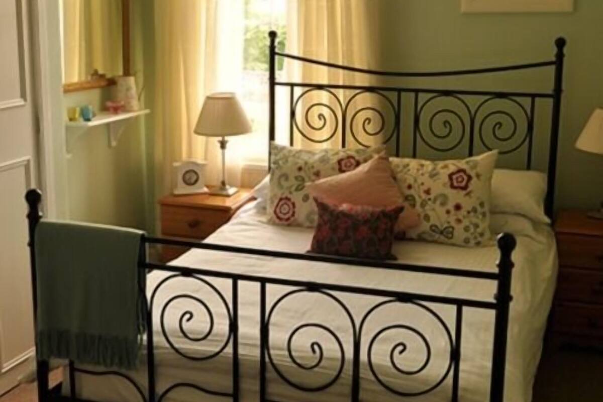 A bedroom with a metal bed frame, a bedside table with a lamp, a chair, and a window with curtains.