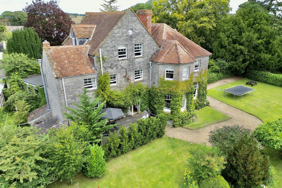 A large stone house with a well-maintained garden, including a lawn, a tennis court, and various shrubs and trees.
