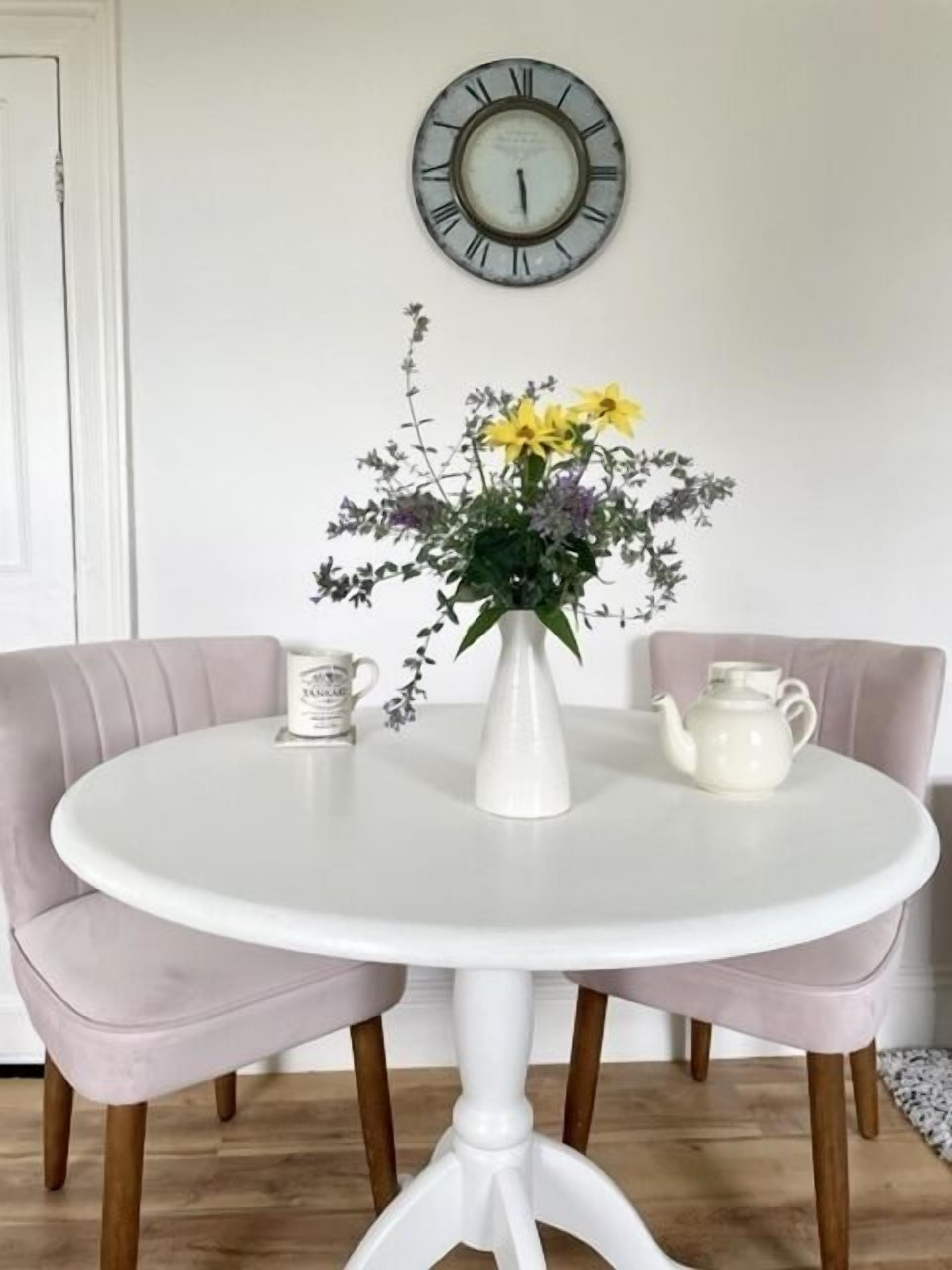 A round white table with a white vase of flowers, two white teapots, and a white mug on a white tablecloth. Two pink upholstered chairs are positioned around the table.