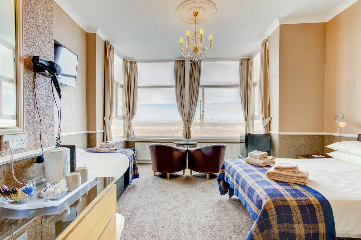 A hotel room with a bed, a desk, two chairs, a chandelier, and a large window with a view.