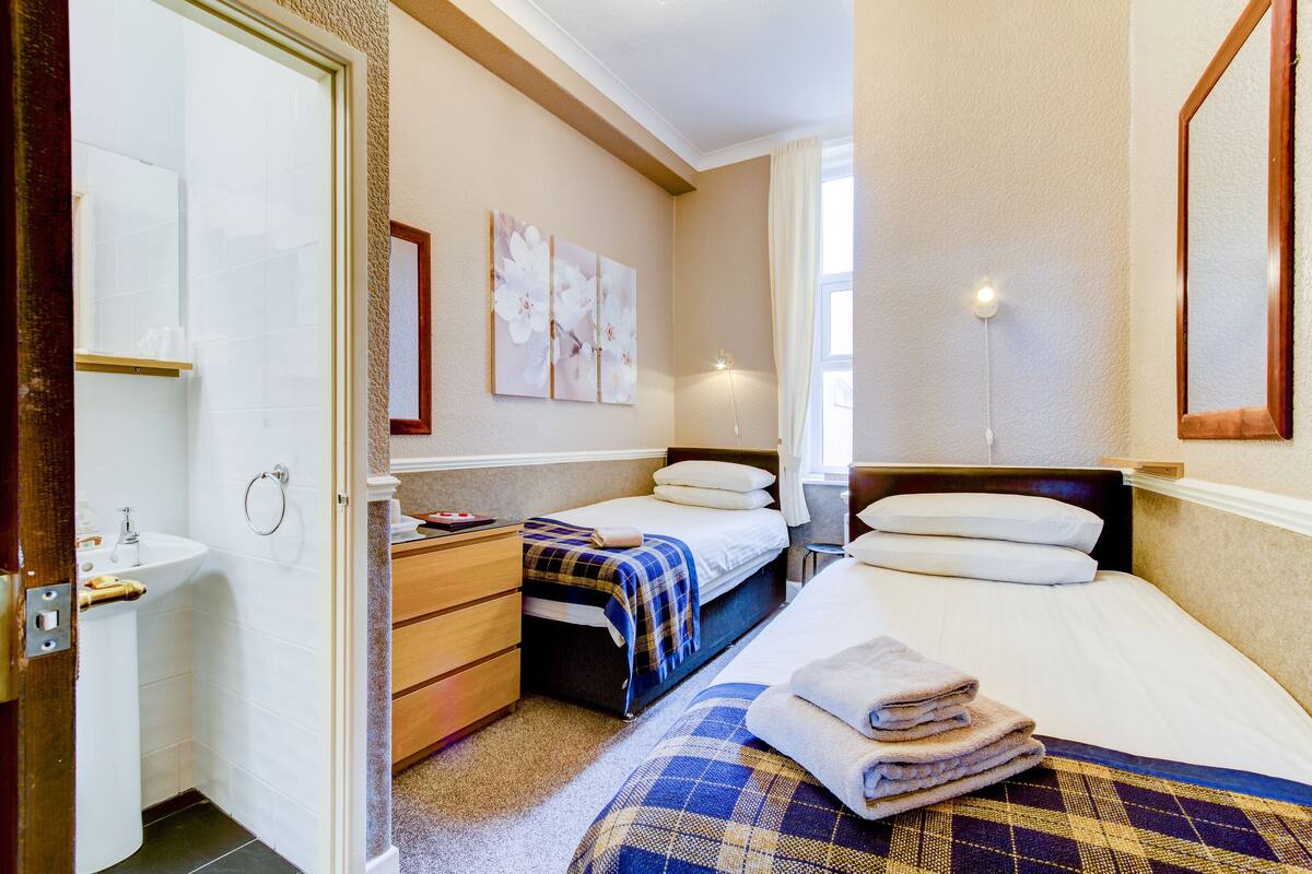 A hotel room with a bed, a bedside table, a small desk, a chair, a window, and a bathroom with a sink and toilet.