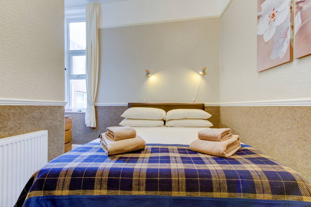 A hotel room with a bed, pillows, a window with curtains, and wall-mounted lights.