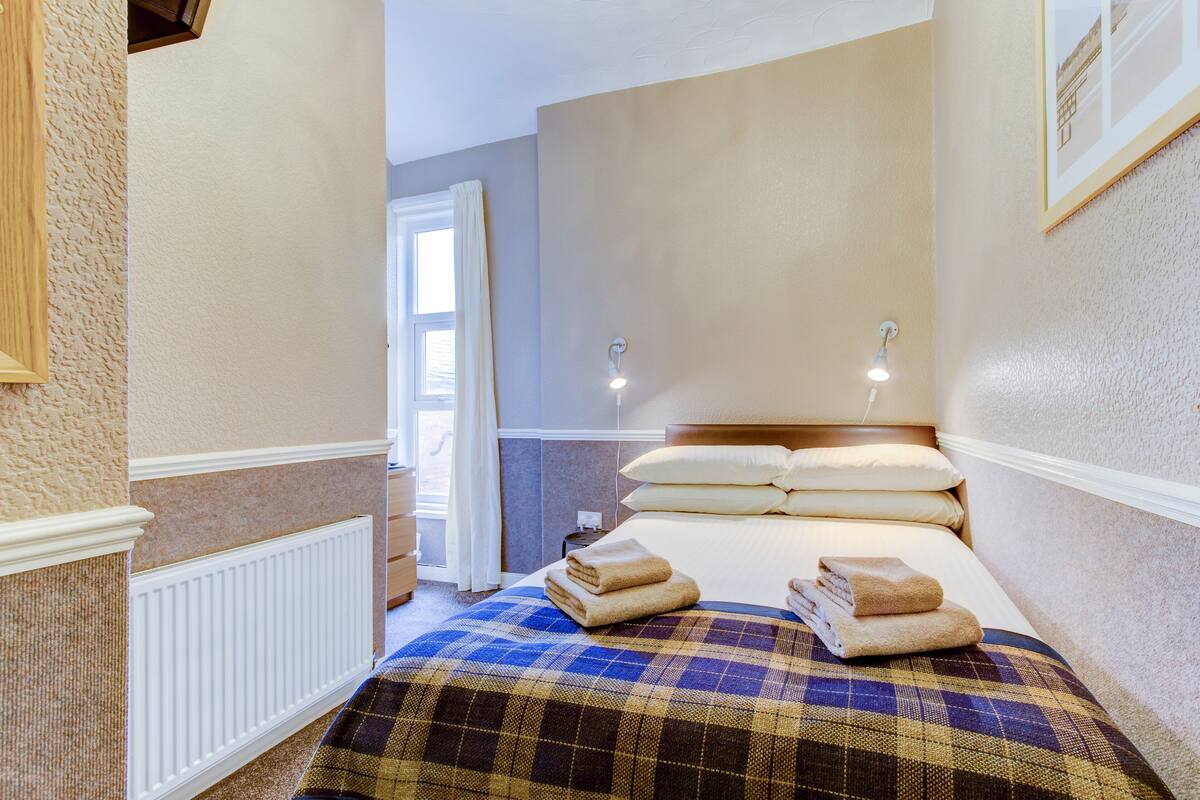 A neatly made bed with a plaid bedspread and multiple pillows, a bedside table with a lamp, and a view of a hallway with a door and window.