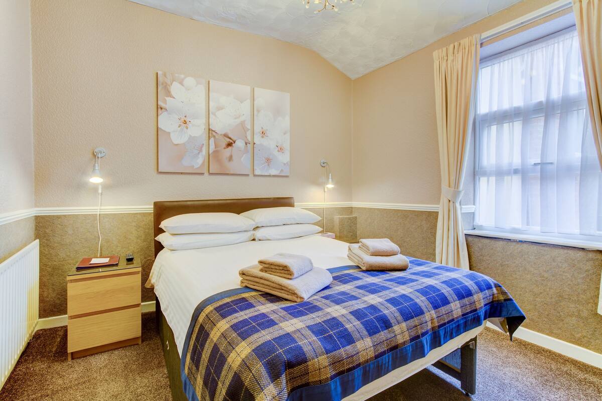 A neatly made bed with a blue and brown checkered bedspread, two bedside lamps, a nightstand with a book and remote, and a window with curtains.