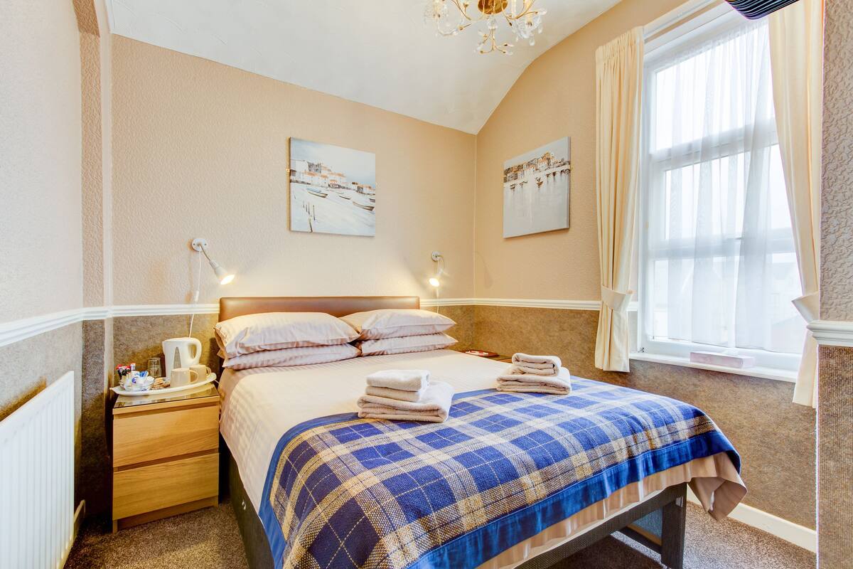 A neatly made bed with a blue and white checkered blanket, a bedside table with a lamp, and two paintings on the wall.