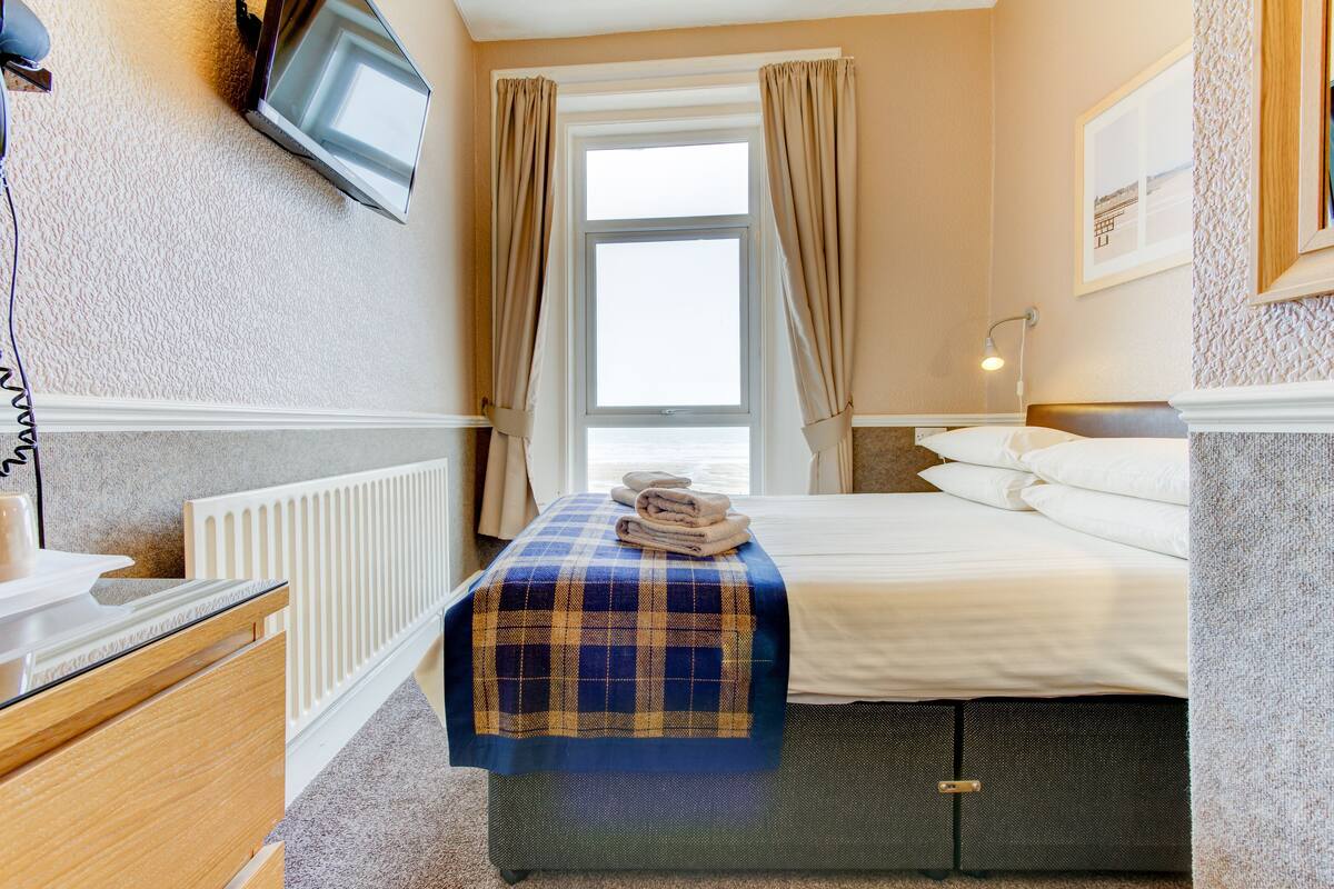 A hotel room with a bed, a bedside table, a TV mounted on the wall, and a window with curtains.