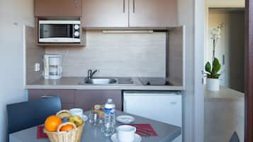 A compact kitchenette with a microwave, coffee maker, and sink. A dining table with a basket of fruit, a water bottle, and a cup is visible.