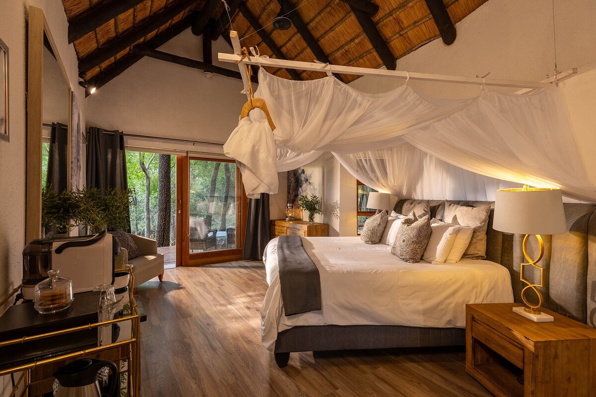 A bedroom with a canopy bed, a wooden desk with a coffee maker, and a view of the outdoors.