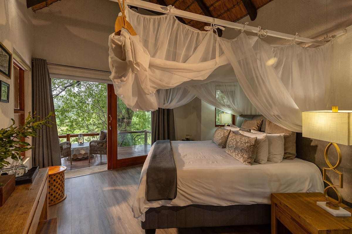 A bedroom with a canopy bed, a wooden floor, a nightstand, a lamp, and a view of the outdoors.