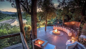 An outdoor dining area with a fire pit, surrounded by tables and chairs, overlooking a river and forested landscape. - Karongwe - Kuname Safari Lodge (Hoedspruit)