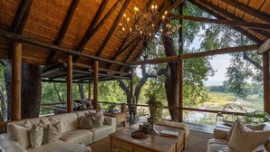A spacious, open-plan living area with a high ceiling and a thatched roof, featuring a large window overlooking a natural landscape. - Karongwe - Kuname Safari Lodge (Hoedspruit)