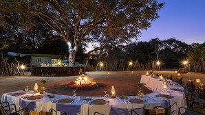 An outdoor dining area with round tables set for a meal, surrounded by trees and a fire pit. - Karongwe - Chisomo Safari Camp (Tzaneen)