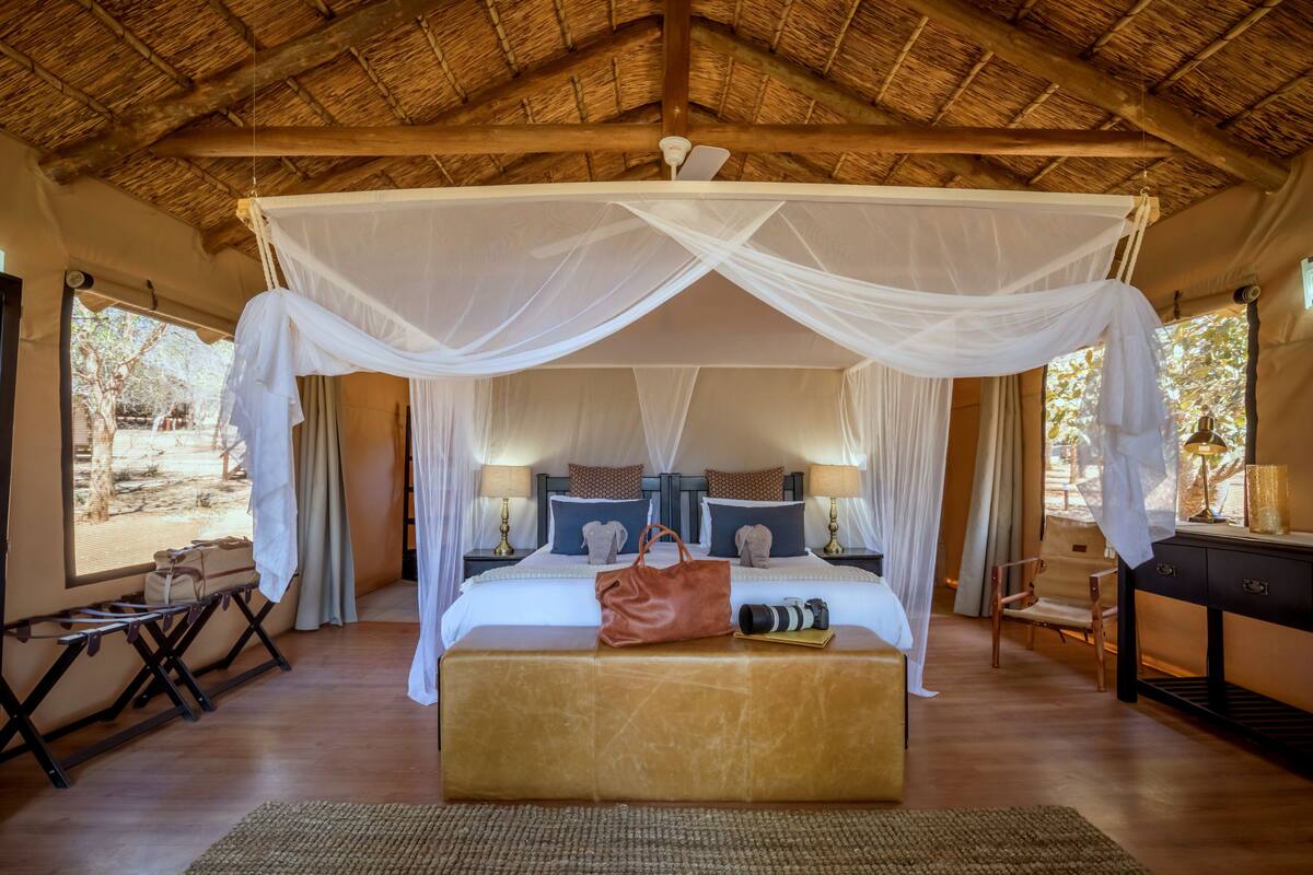 A room with a bed under a canopy, bedside tables with lamps, a bench, and a view of the outdoors.