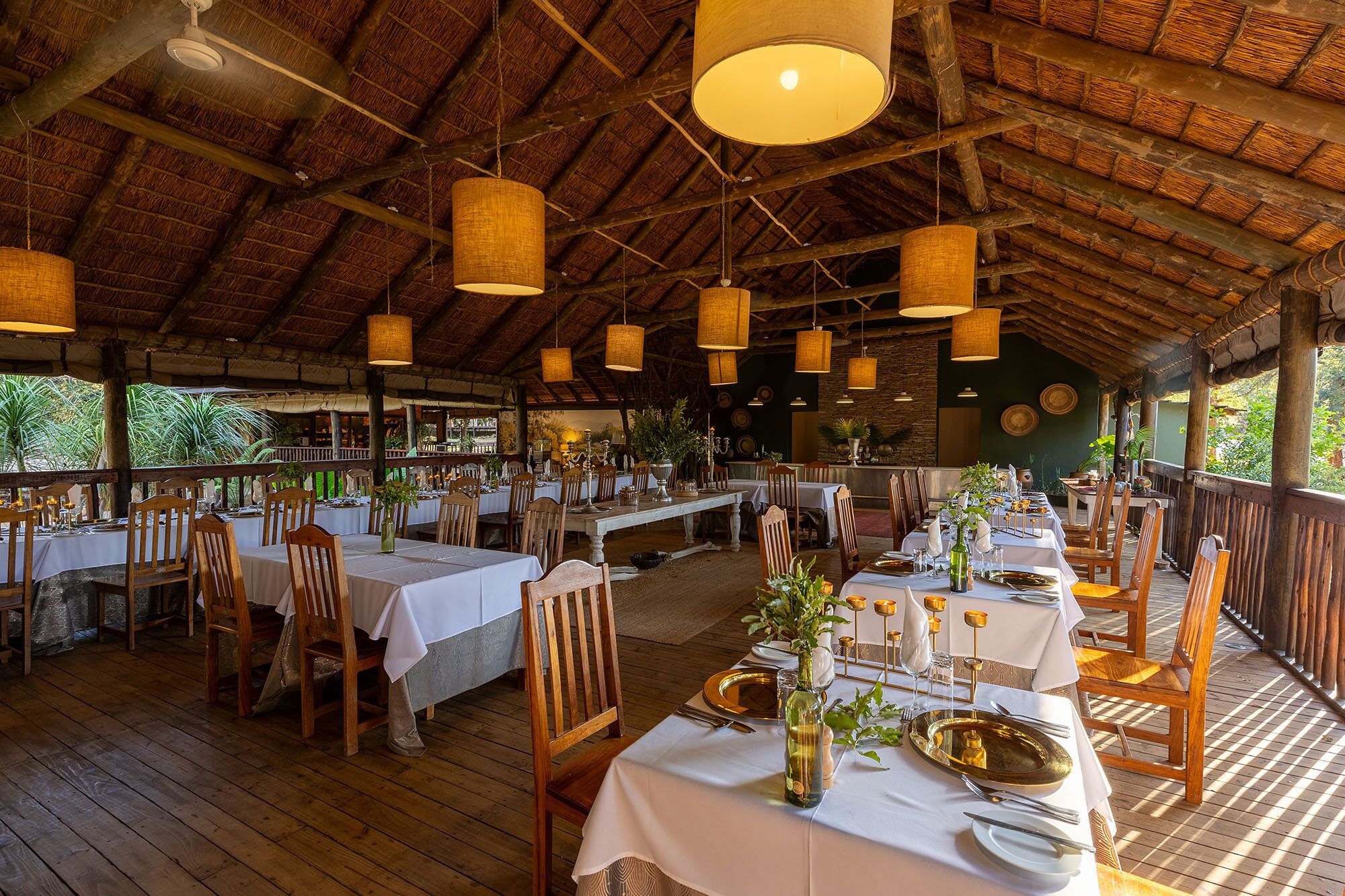 An outdoor dining area with wooden tables and chairs, thatched roof, and hanging light fixtures.