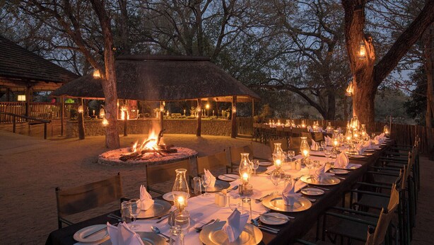 An outdoor dining area with a long table set for a meal, surrounded by trees and a thatched-roof structure. - Karongwe - Chisomo Safari Camp (Tzaneen)