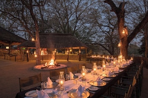 An outdoor dining area with a long table set for a meal, surrounded by trees and a thatched-roof structure. - Karongwe - Chisomo Safari Camp (Tzaneen)