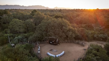 An aerial view of a campsite with a circular seating area, surrounded by dense forest and mountains in the background.