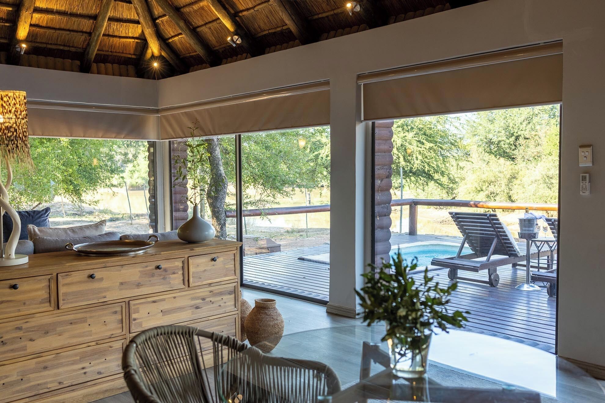 Una habitación amplia con un ventanal, un aparador de madera, una mesa de vidrio y vistas a una terraza exterior con sillones de descanso y piscina.